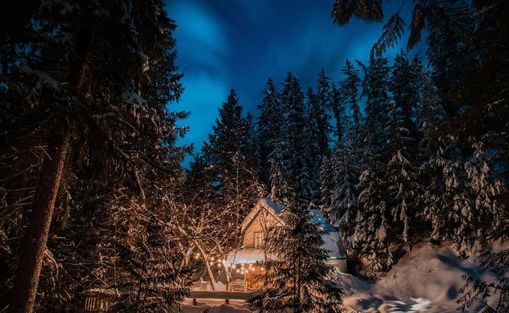 Fotografia di una baita in mezzo al bosco con il cielo "nell'ora blu" e alcune luci accede sul patio della casa